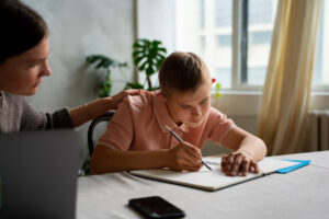A child in a pink shirt writes in a notebook during a study session, while an adult offers support from beside them.