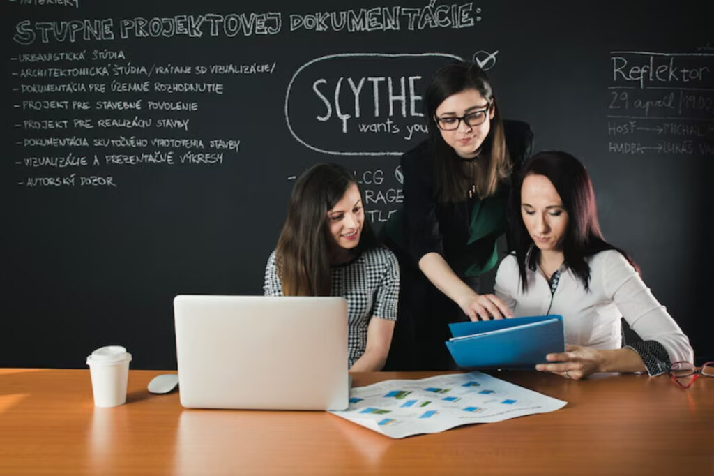 Three women collaborate at a desk with a laptop, papers, and a coffee cup, discussing project documentation against a chalkboard backdrop.