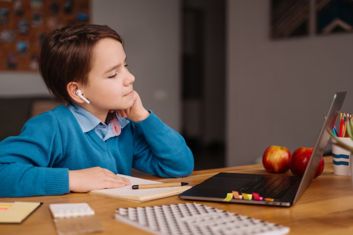 A child in a blue sweater sits at a desk with a laptop, notepad, apples, and colored pencils, appearing deep in thought.
