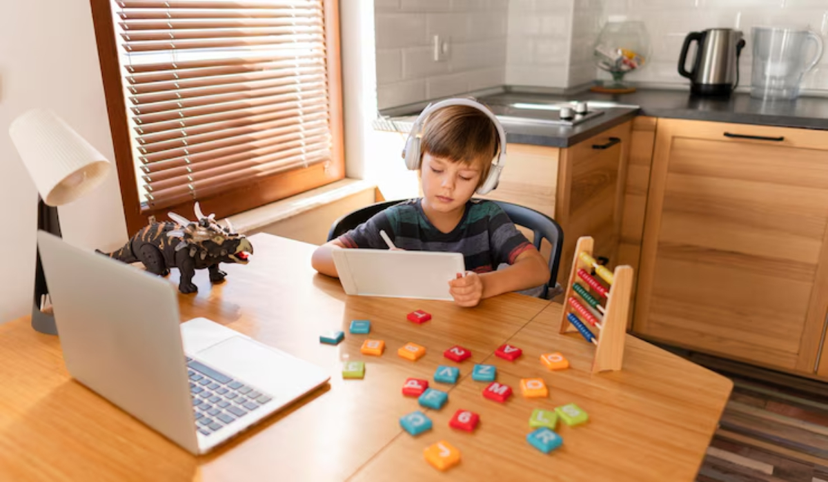 A child wearing headphones sits at a desk with a tablet, surrounded by colorful letter tiles and a toy dinosaur in a bright kitchen.