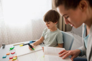 A child sits at a table drawing in a notebook, surrounded by colorful letters and markers, while an adult reads from a book.