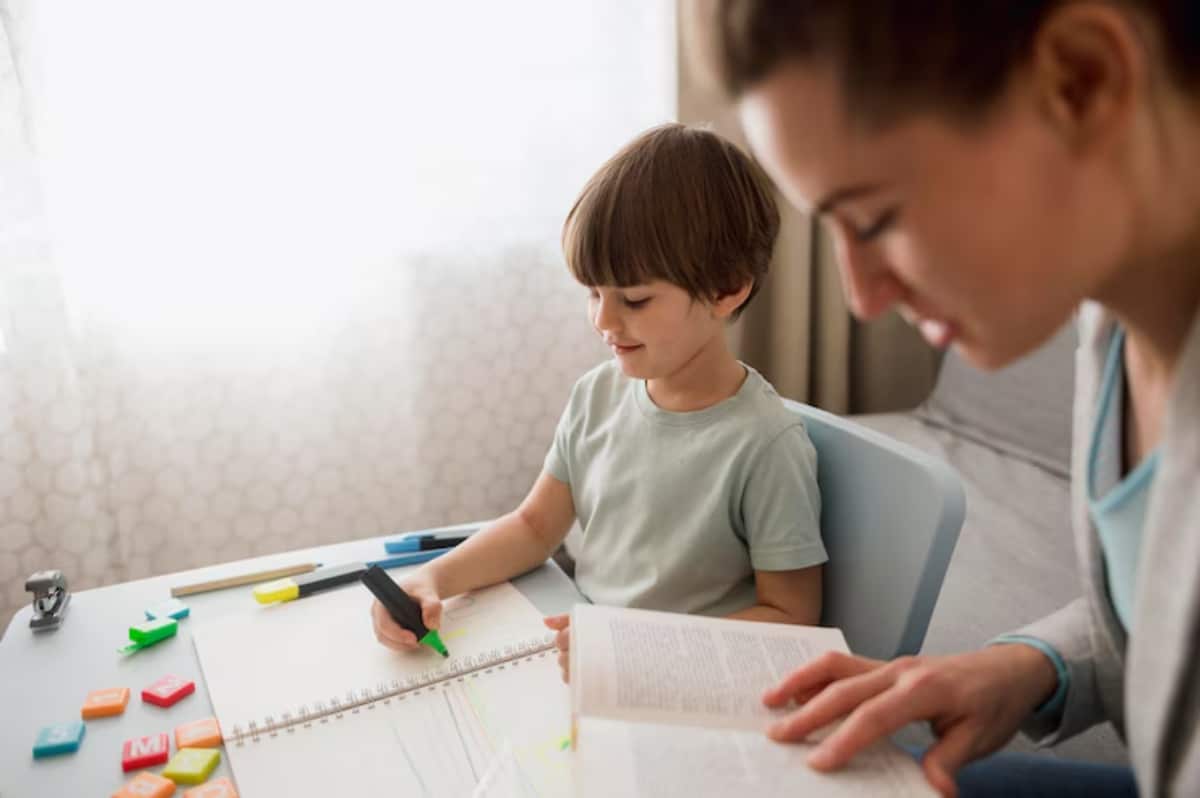 A child sits at a table drawing in a notebook, surrounded by colorful letters and markers, while an adult reads from a book.