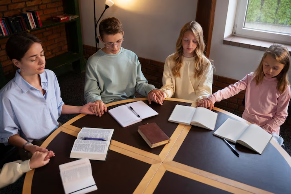 A group of four individuals holding hands around a table with open notebooks and a book, in a cozy, well-lit reading space.
