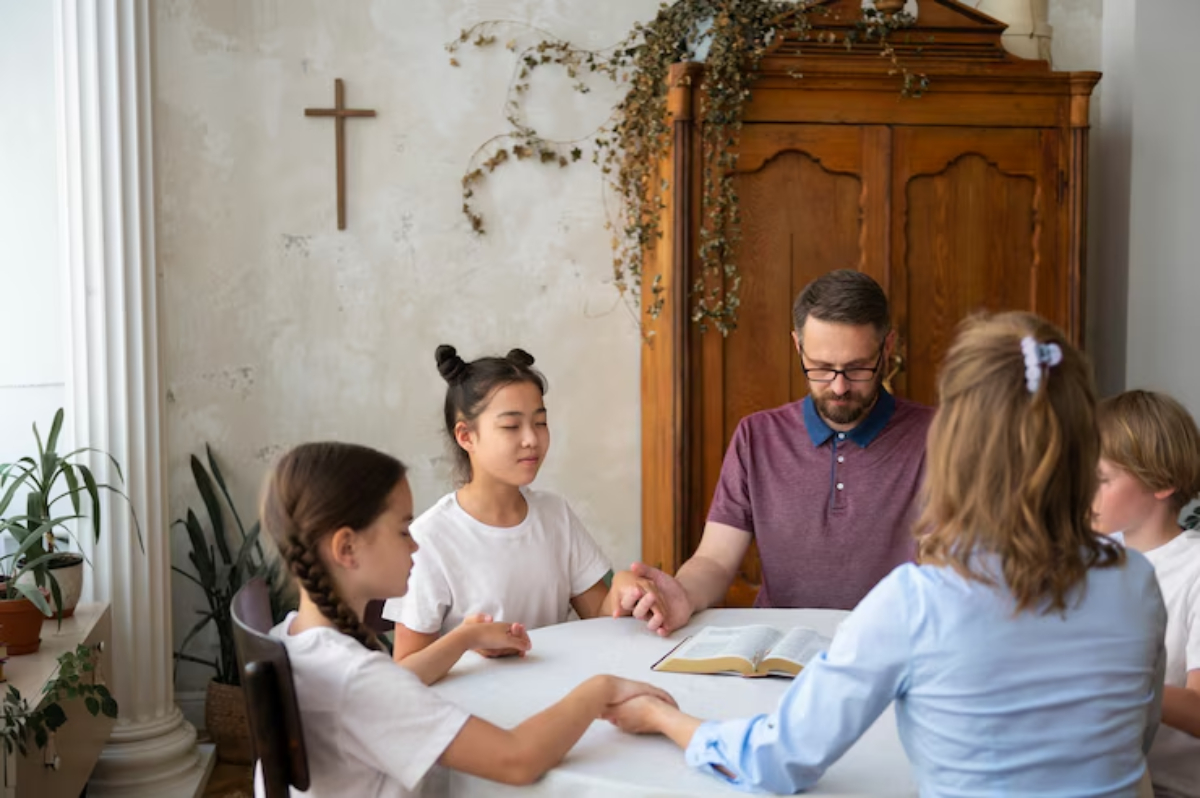 A group of people hold hands in prayer around a table, with an open Bible in front, in a warmly decorated indoor space.