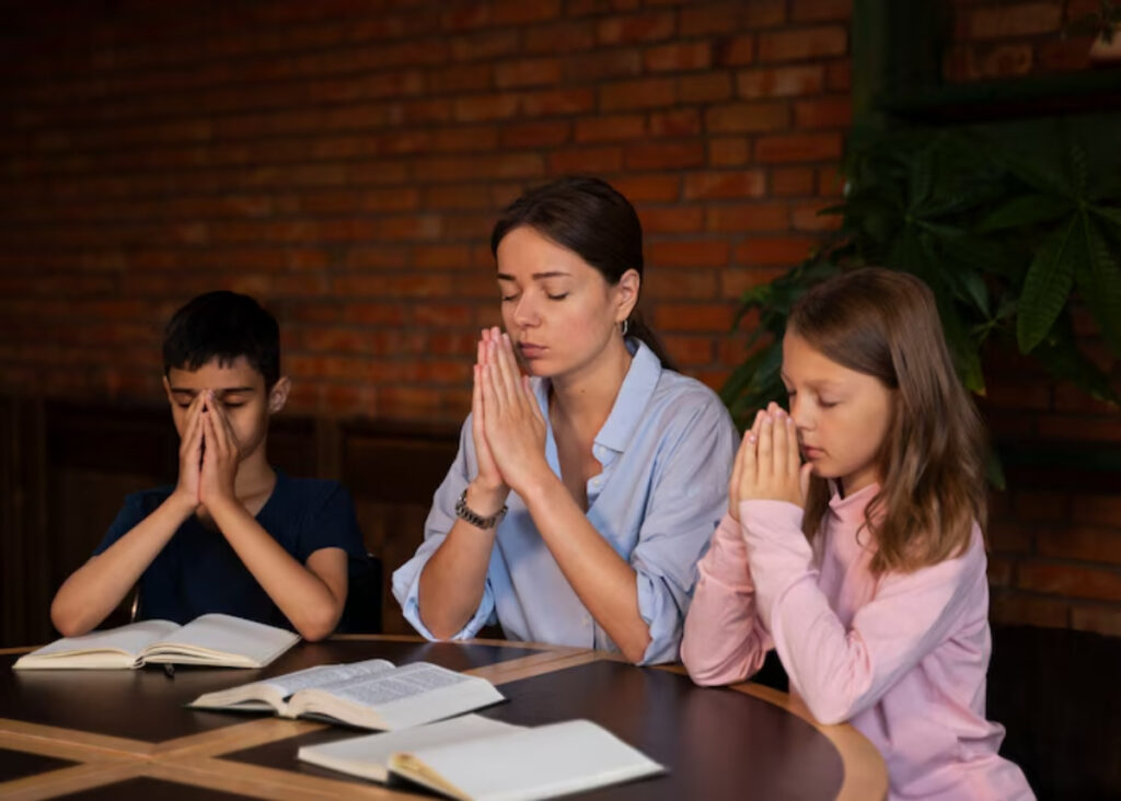 A woman and two children sit at a table, hands clasped in prayer, with open books in front of them against a rustic brick background.