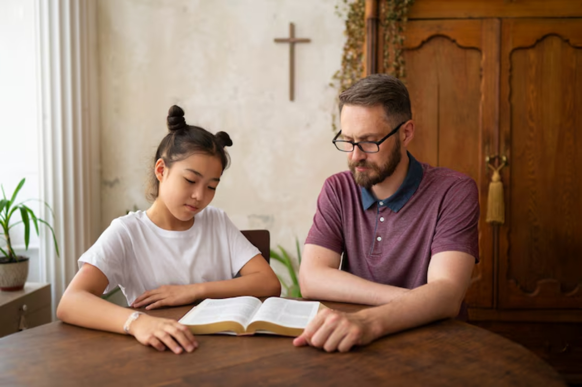 A child and an adult are seated at a wooden table, engaged in reading a book, with a cross on the wall and plants in the background.