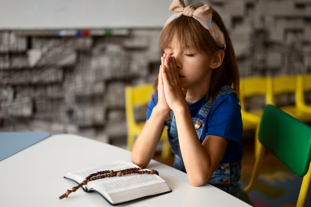 A young girl sits at a table with a Bible open in front of her, clasping her hands in prayer, surrounded by colorful chairs.