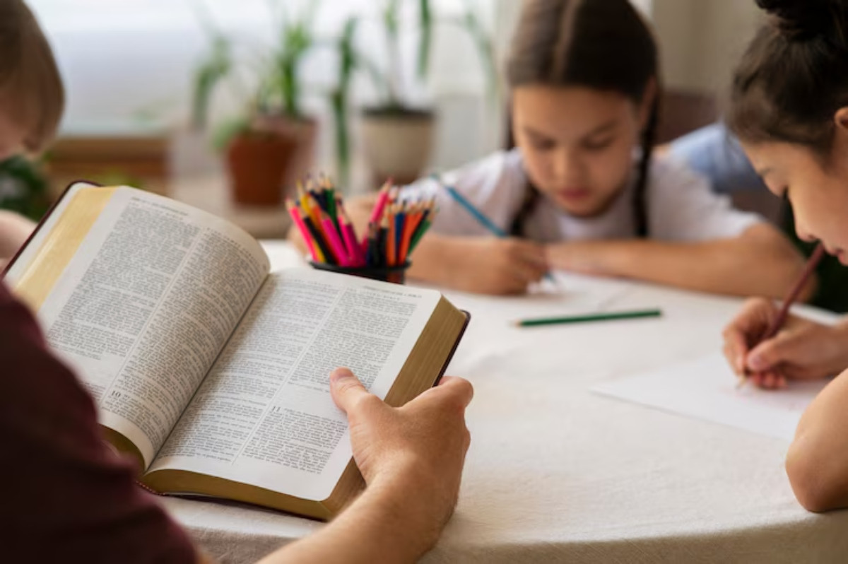 A person is reading a book while two children engage in drawing on a table, surrounded by colorful pencils and plants.