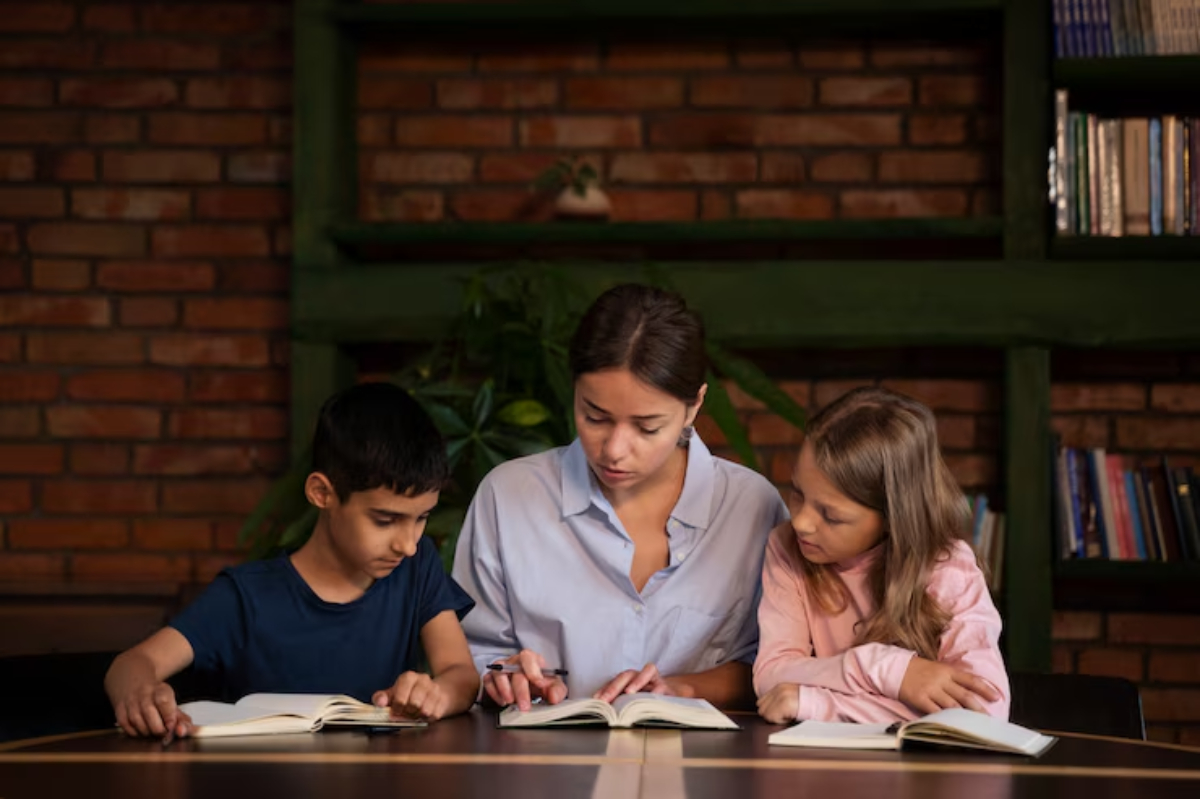 A woman sits at a table with two children, all engaged in reading books in a cozy, rustic setting with brick walls and bookshelves.