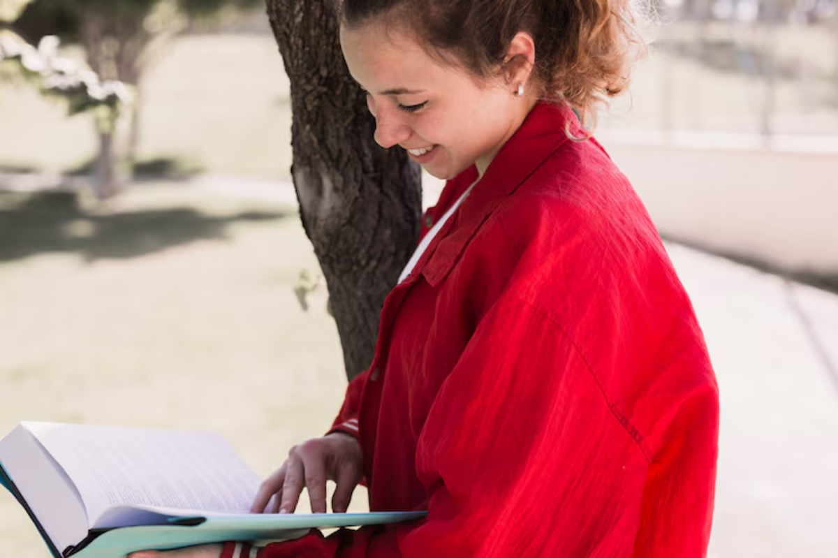 A person in a red jacket stands by a tree, engrossed in reading a large book on a sunny day in a park setting.