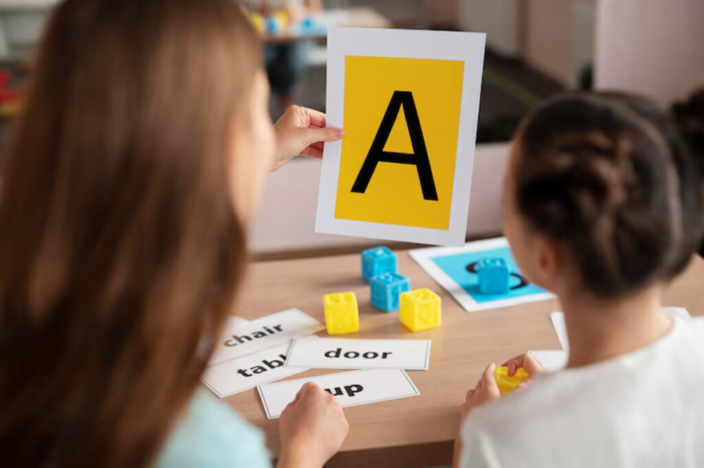 A child shows a letter A card while sitting at a table with word cards and colorful building blocks in a learning environment.