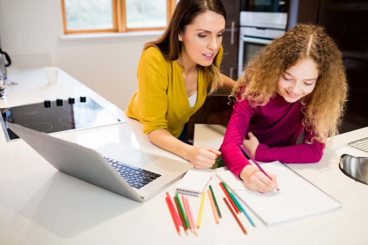 A woman guides a girl in a colorful top as they work on a drawing at a kitchen table surrounded by pencils and a laptop.