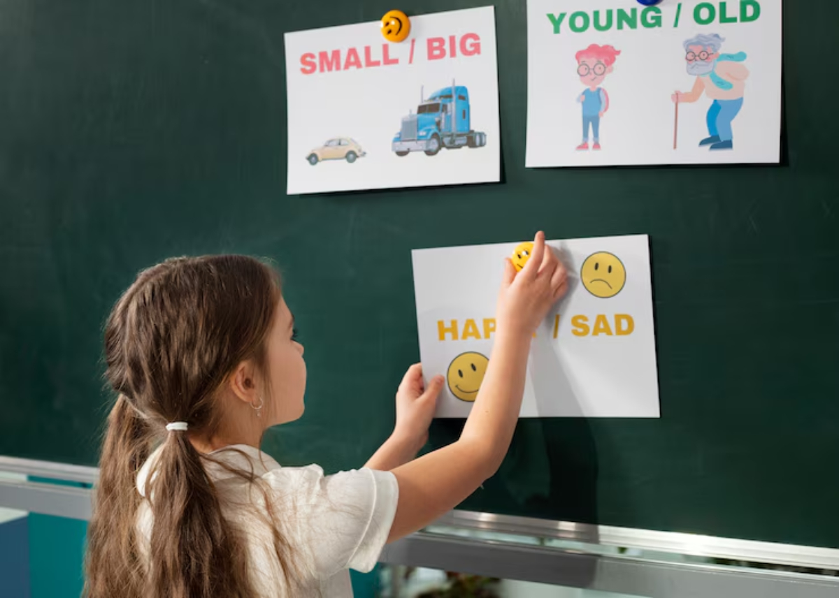 A child places a sticker on a chart displaying emotions, with signs for HAPPY / SAD, SMALL / BIG, and YOUNG / OLD on a chalkboard.