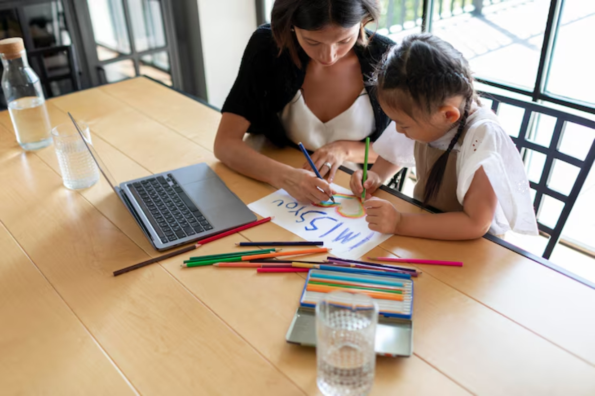 A woman and a child draw together at a wooden table, surrounded by colorful markers and a laptop, with a glass of water nearby.