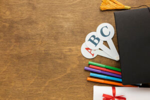A black graduation cap, colorful markers, and ABC flashcards are arranged on a brown wooden surface, alongside a gift-wrapped diploma.