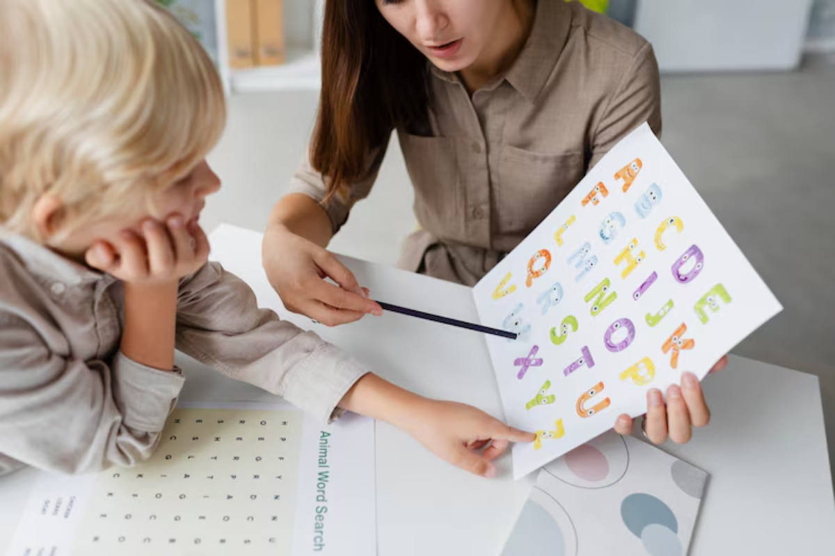A woman teaches a child letters with a colorful alphabet chart at a table, surrounded by educational materials and worksheets.