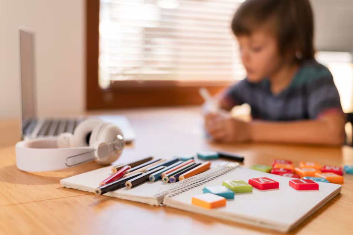 A wooden table with colorful stationery, a notepad, and headphones, next to a laptop, in a bright, sunny room setting.