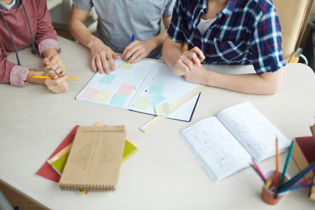 Three students collaborate at a table filled with stationery, notebooks, and sticky notes, focusing on a shared project.
