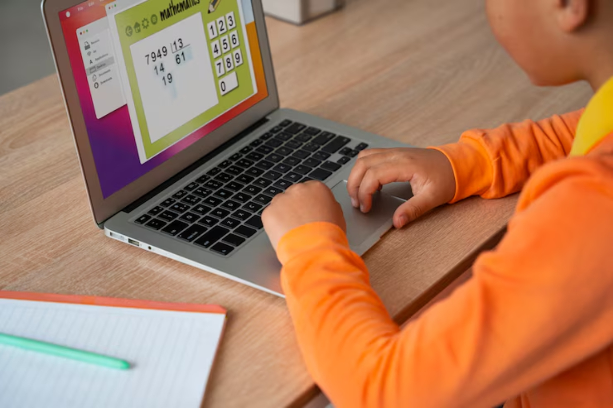 A child in an orange shirt interacts with a laptop displaying a math game, while a notebook lies open nearby.