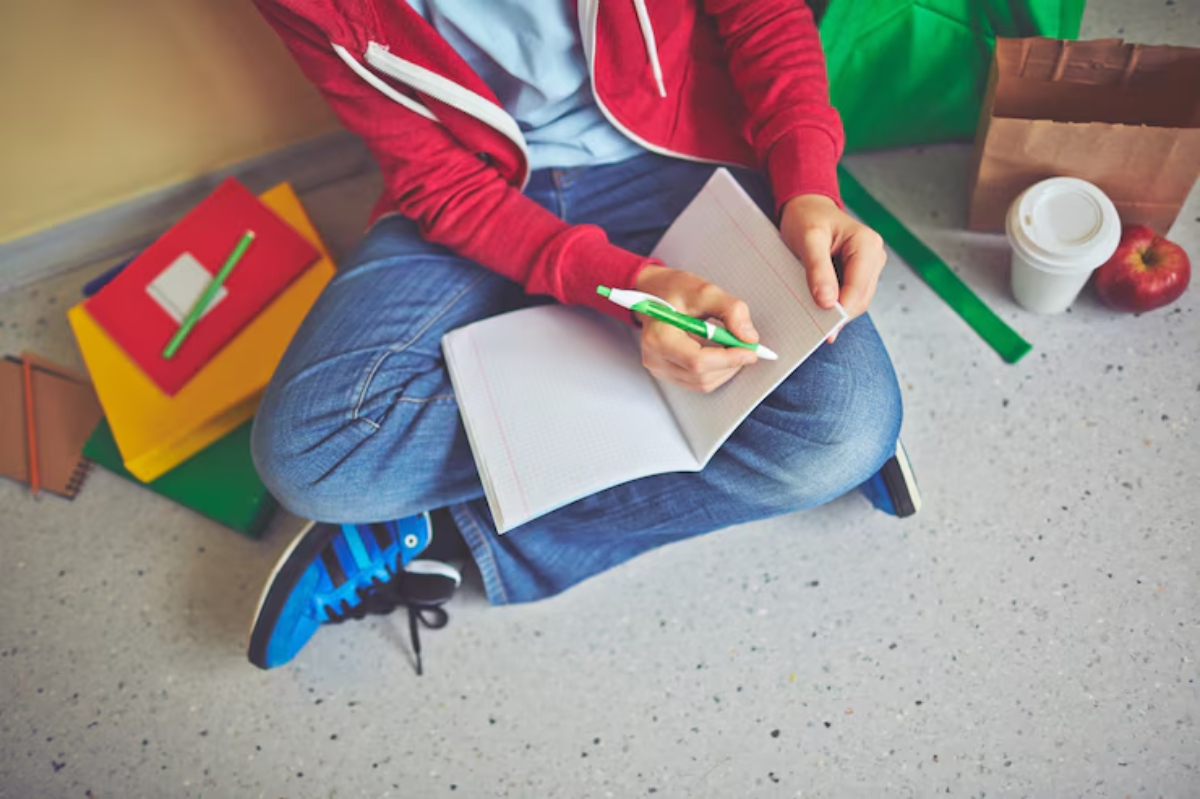 A person sits on the floor, writing in a notebook with a green pen, surrounded by colorful stationery, a drink, and an apple.