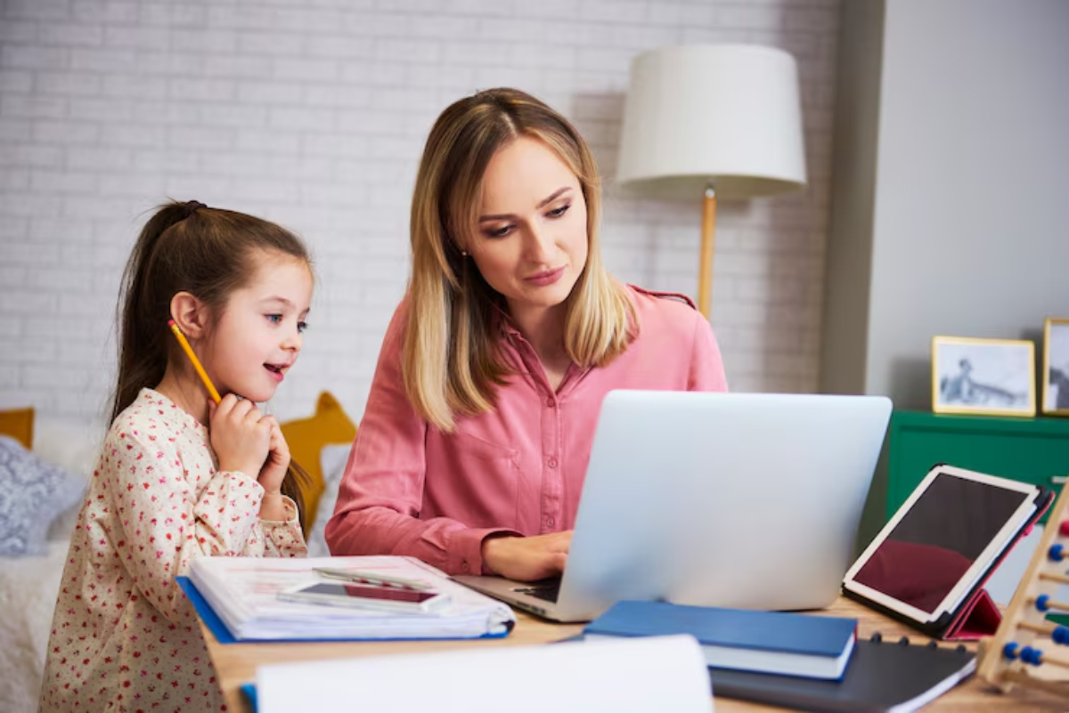 A woman in a pink shirt assists a child with a pencil, both engaged in a laptop activity at a cozy home workspace.