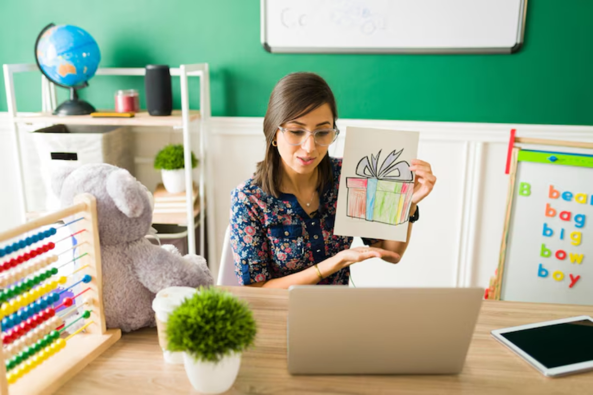 A person holds a colorful drawing of a gift while seated at a desk with a laptop, educational toys, and a chalkboard in a bright classroom.