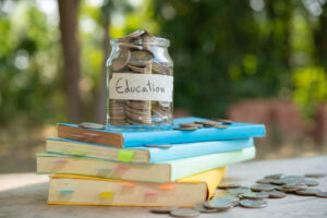 A jar labeled Education filled with coins sits atop colorful stacked books, surrounded by scattered coins on a wooden table outdoors.