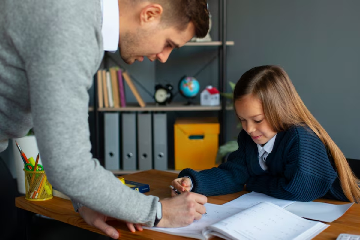A teacher assists a young student at a wooden desk, with notebooks and stationery, in a bright, modern classroom setting.