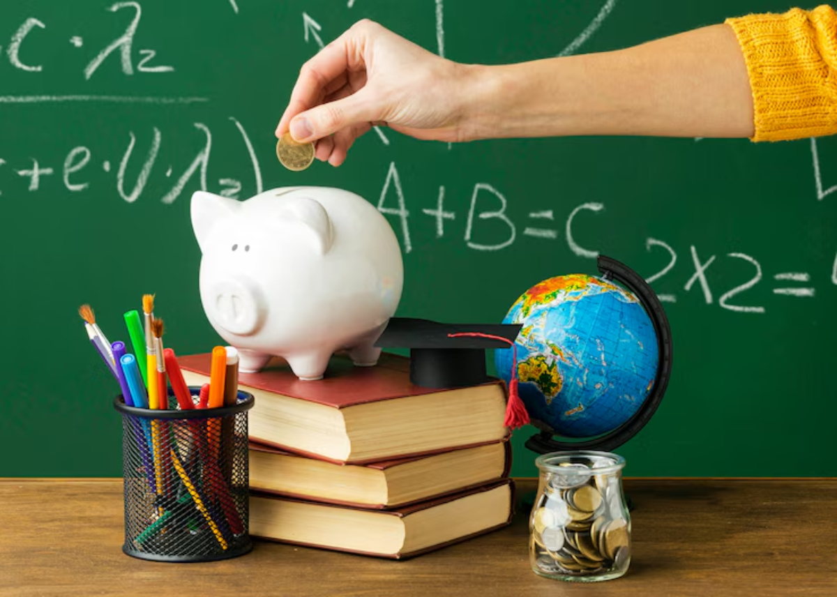 A hand drops a coin into a piggy bank atop books, alongside a globe, a jar of coins, and colorful pens on a classroom table.