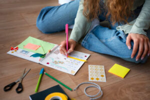 A person with curly hair writes in a decorated notebook on the floor, surrounded by stickers, scissors, and stationery supplies.