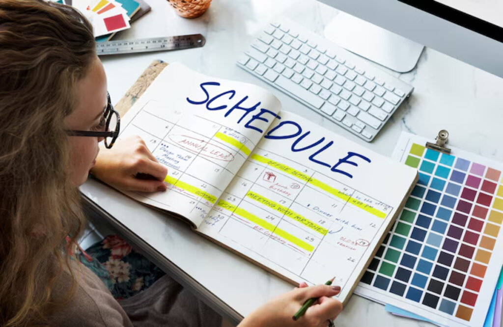 A person writes in a planner marked SCHEDULE, surrounded by color palettes and a computer keyboard on a desk.