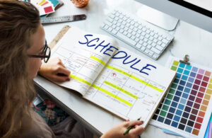 A person writes in a planner marked SCHEDULE, surrounded by color palettes and a computer keyboard on a desk.