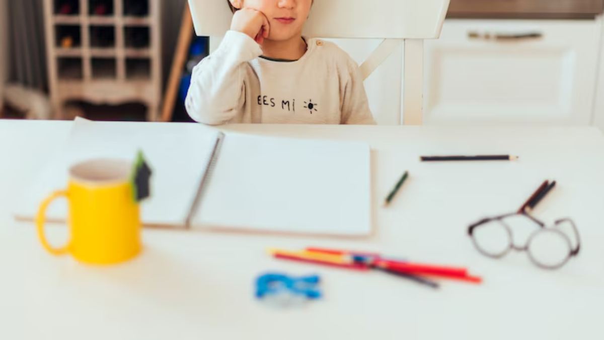 A young boy sits at a table, resting his chin on his hand, with art supplies and a blank notebook in front of him, deep in thought.