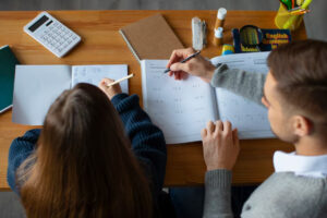 A study session at a wooden table with notebooks, a calculator, and writing materials as two people work together.