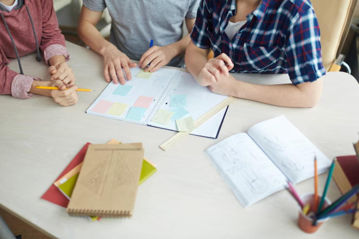 Three students collaborate around a table, using notebooks, sticky notes, and stationery to brainstorm ideas for a project.