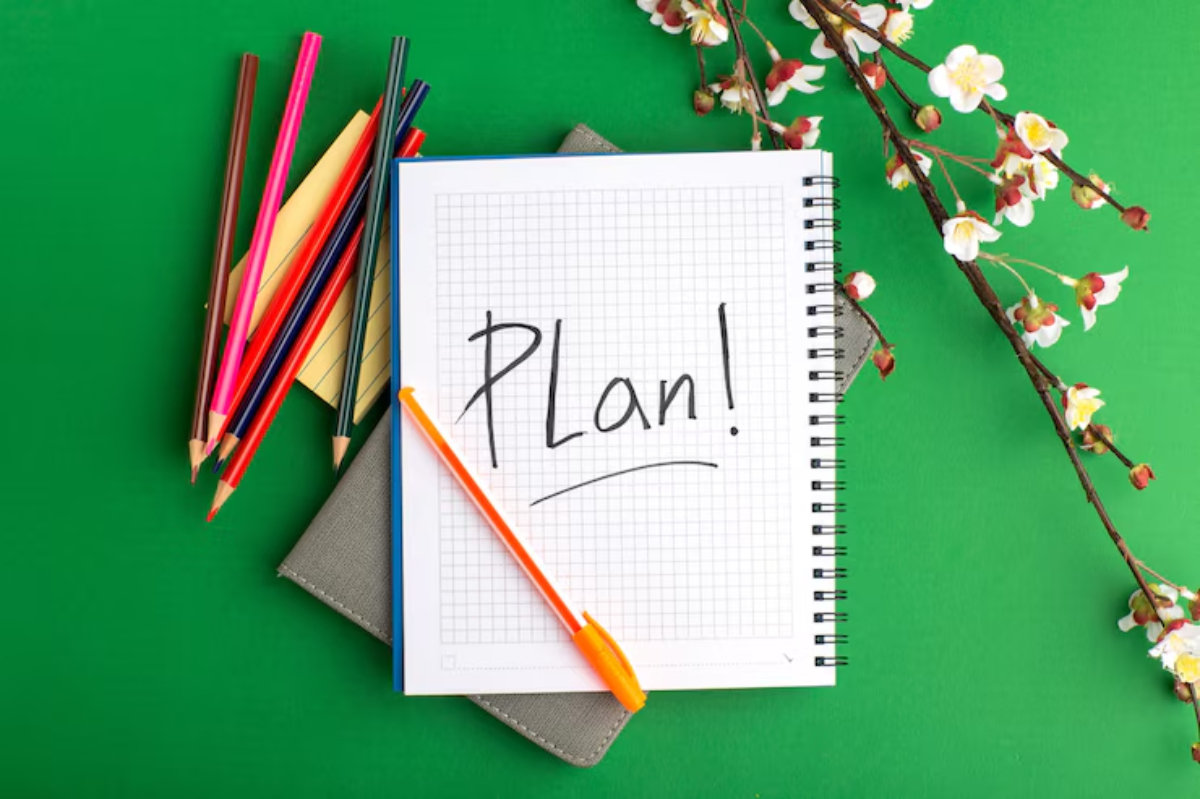 A notebook with Plan! written on a grid page, surrounded by colorful pens, paper, and a branch of flowers on a green background.