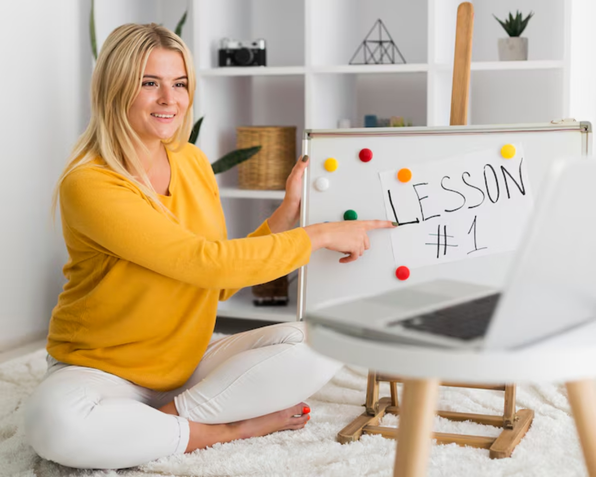 A person in a yellow sweater sits cross-legged, pointing at a whiteboard labeled LESSON #1, with a laptop nearby.