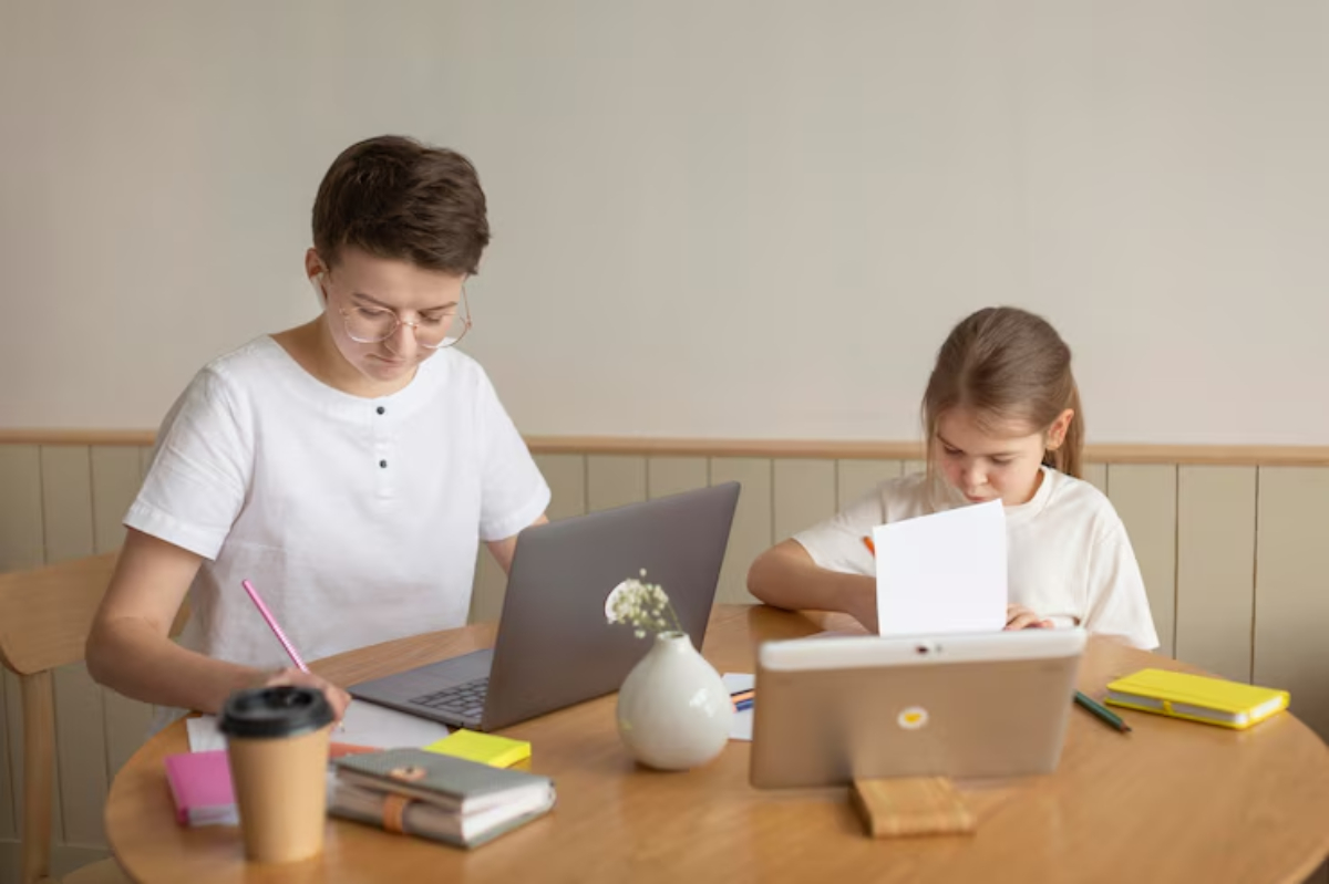 A young adult and child are seated at a table, working on laptops and writing, surrounded by stationery and a coffee cup.