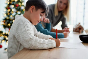 A child in a cozy white sweater writes with a red pencil at a table, while two adults engage in similar activities, festive decorations in view.