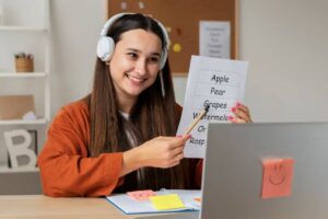 A person wearing headphones holds a sheet listing fruits while seated at a desk with a laptop and colorful sticky notes.