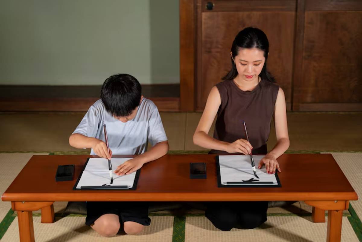 A child and an adult practice calligraphy at a low wooden table, focused on their ink and paper in a serene indoor setting.