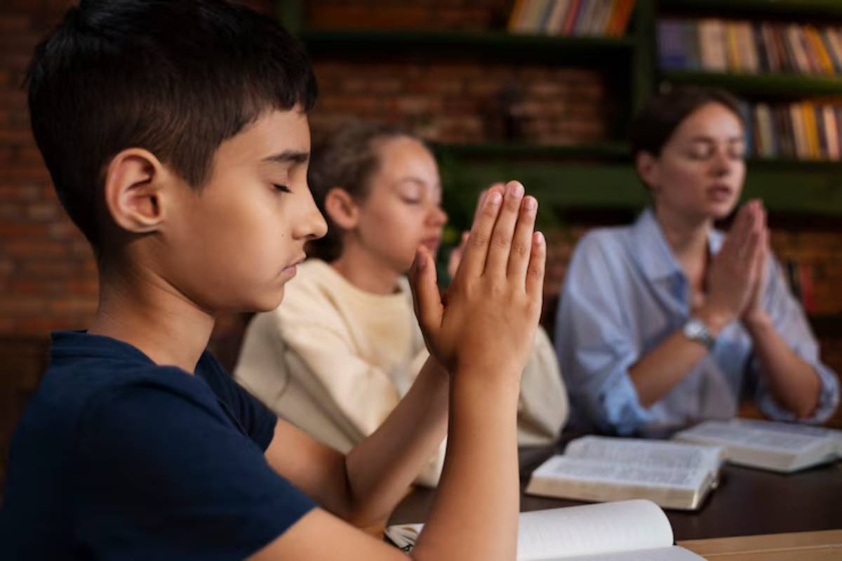 A group of individuals sits at a table with books, hands clasped in prayer against a backdrop of brick and wooden shelves.