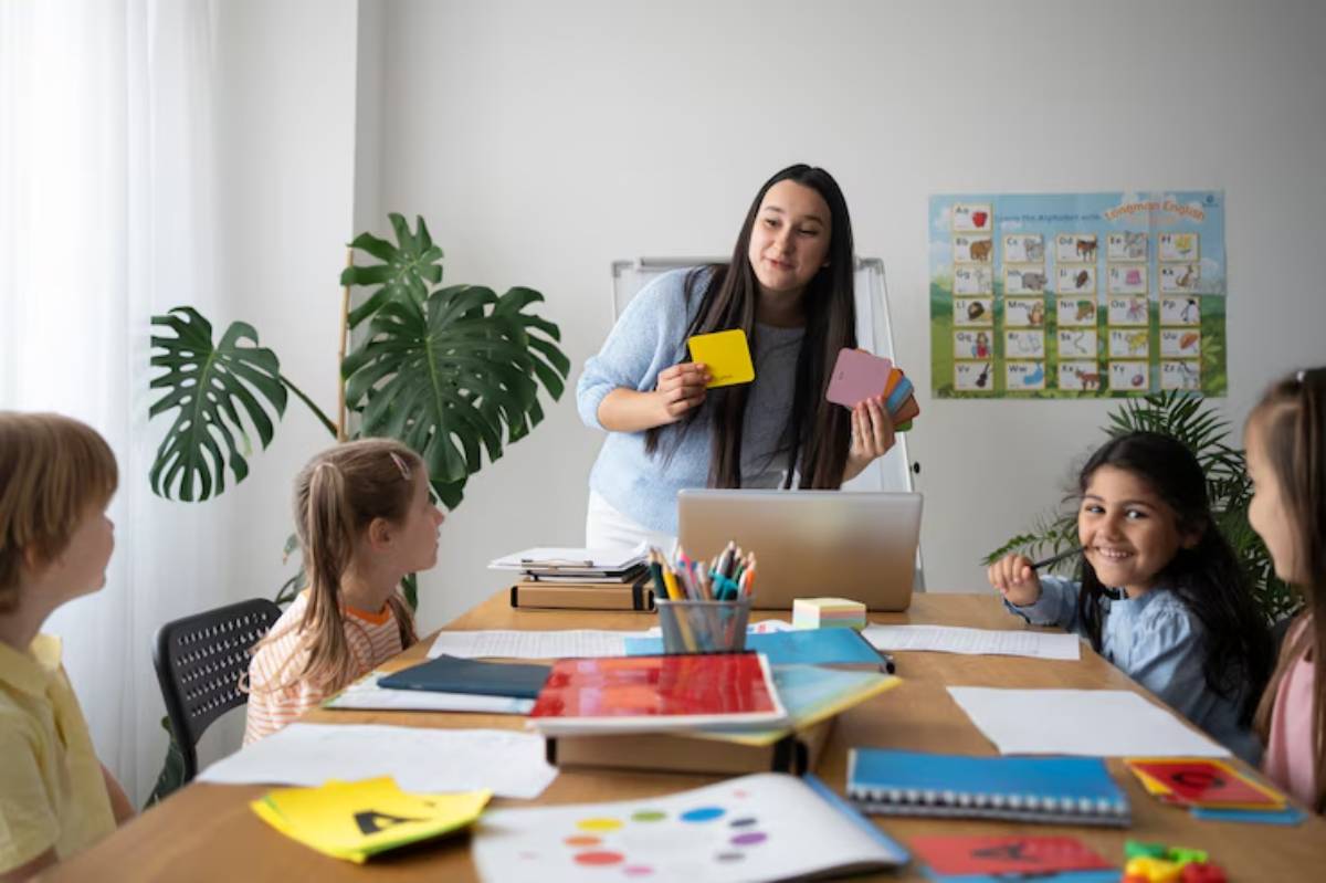 A teacher engages with children in a classroom, using colorful cards, surrounded by educational materials and plants.