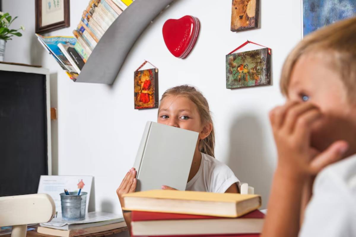 A child sits at a table with stacked colorful books, holding an open, blank book, surrounded by art on the wall and a plant nearby.