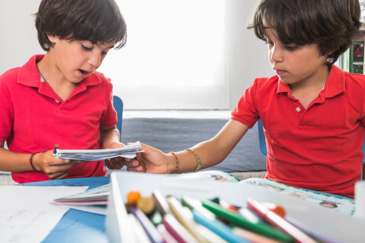Two children in red shirts engage in a study session, exchanging notebooks over a table filled with school supplies and art materials.