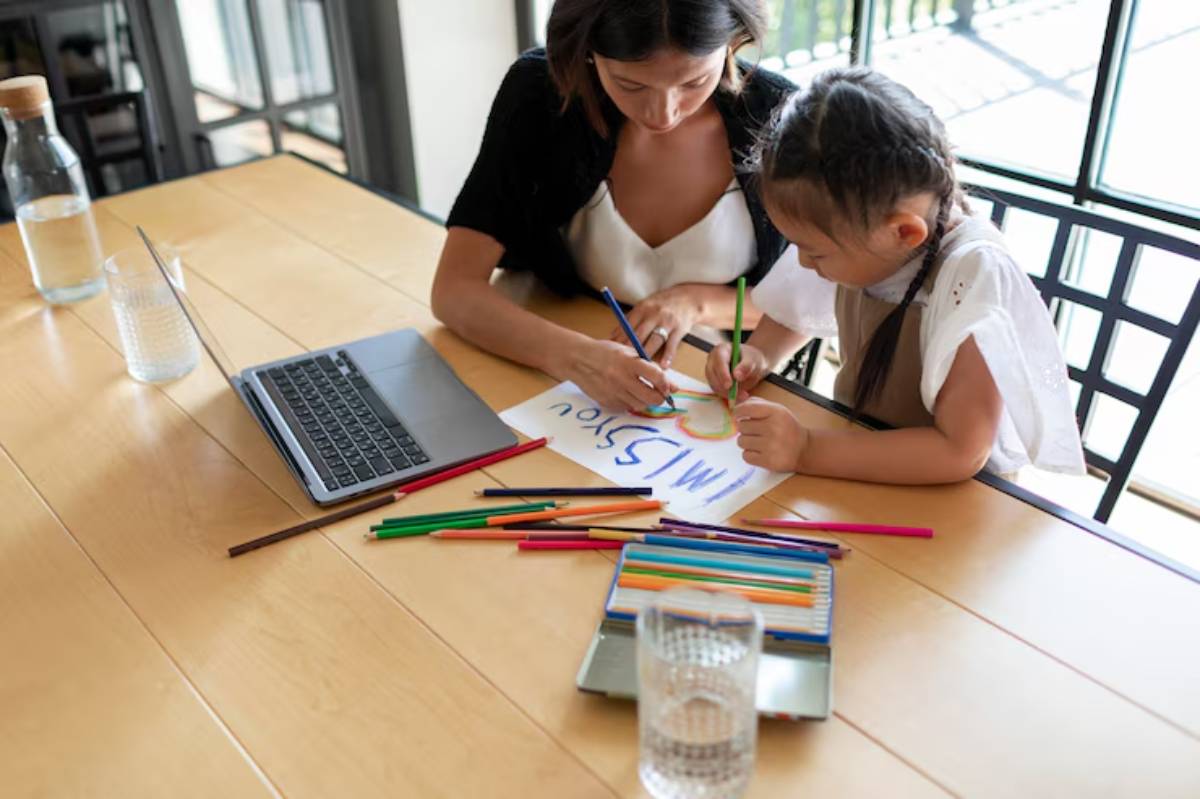 A woman and a girl work together at a table, using colorful markers to create art on a large sheet of paper, with a laptop nearby.