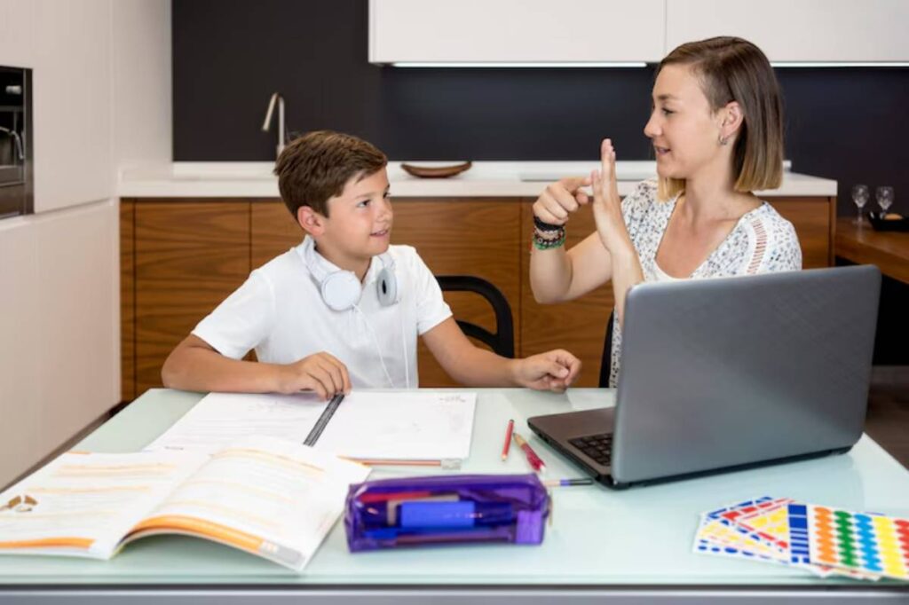 A woman teaches a boy at a glass table covered with notebooks, pens, and a laptop in a modern kitchen setting.