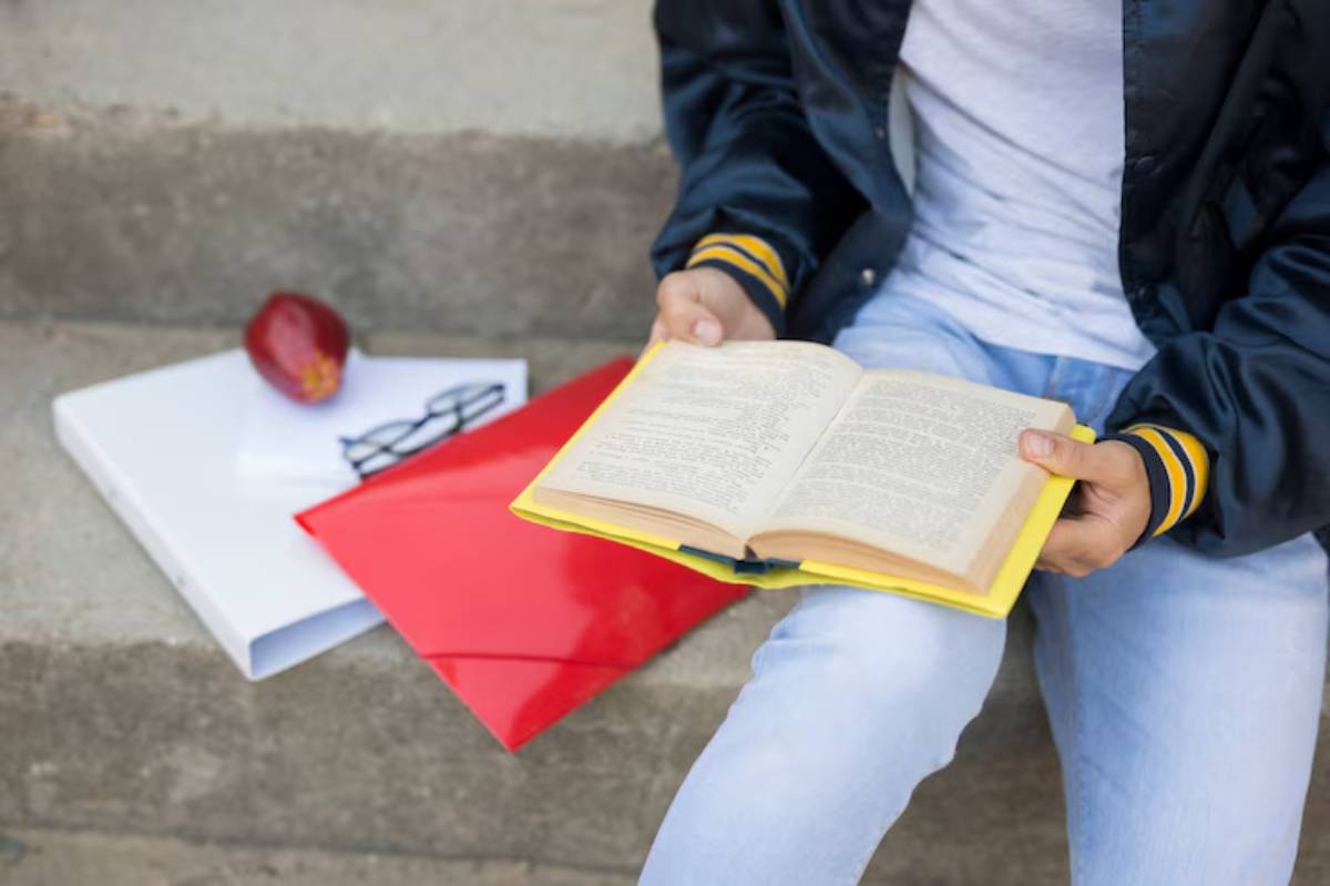 A person reading a large book while sitting on steps, with a red folder, white binder, glasses, and an apple nearby.