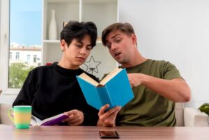 Two individuals sit at a table, focused on a blue book while one points at the pages, surrounded by cozy, modern decor.