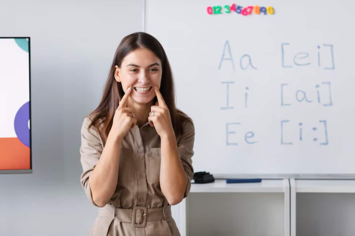 A woman in a beige outfit stands in front of a whiteboard with letters and phonetic symbols, preparing to teach.